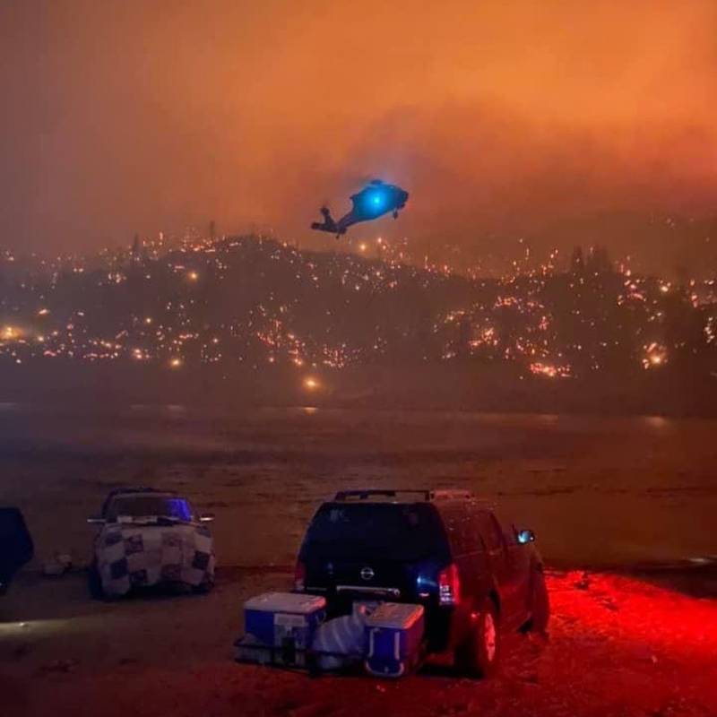 A helicopter hovers over the lake with two trucks on the shore and a hillside burning beyond. Dark sky is completely orange and smoke-filled.