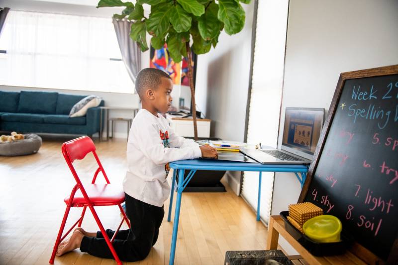 A young boy kneels at a desk in his home to use a laptop computer.