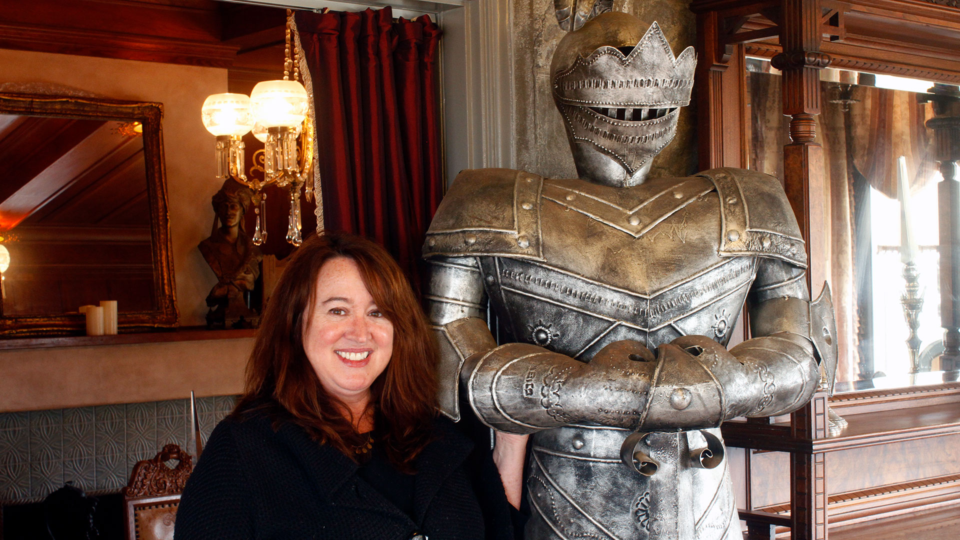 A woman stands next to a large suit of armor in a room full of dark wood.