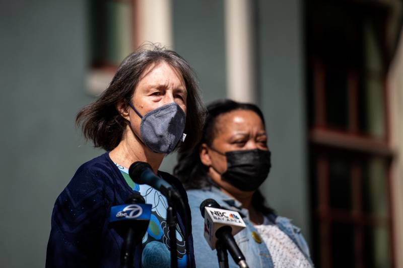 Senator Nancy Skinner, wearing a face mask and standing in front of microphones, looks into the distance.