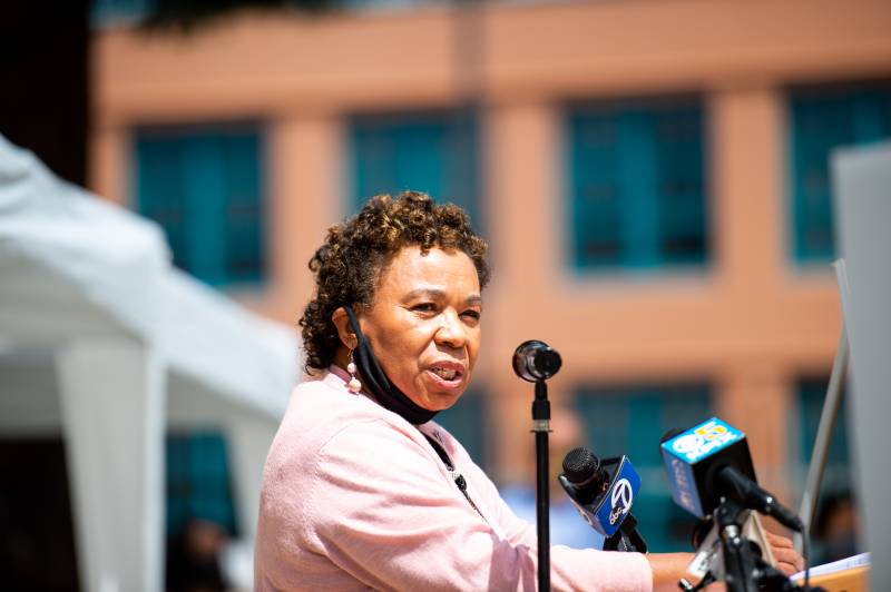 Barbara Lee speaks in front of news microphones, wearing a light pink shirt with a mask under her chin.