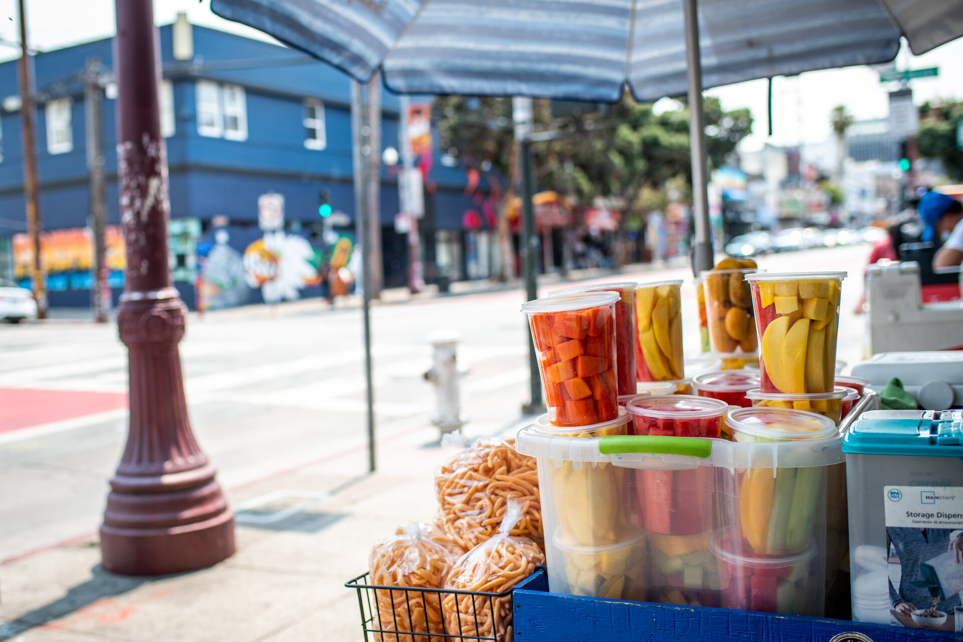Clear plastic tubs of brightly colored fruit beneath an awning, with a city block in the background.