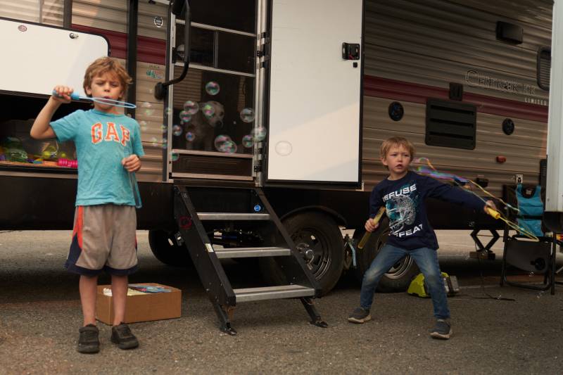 Two boys blow bubbles through long wands in front of the stairway to an RV