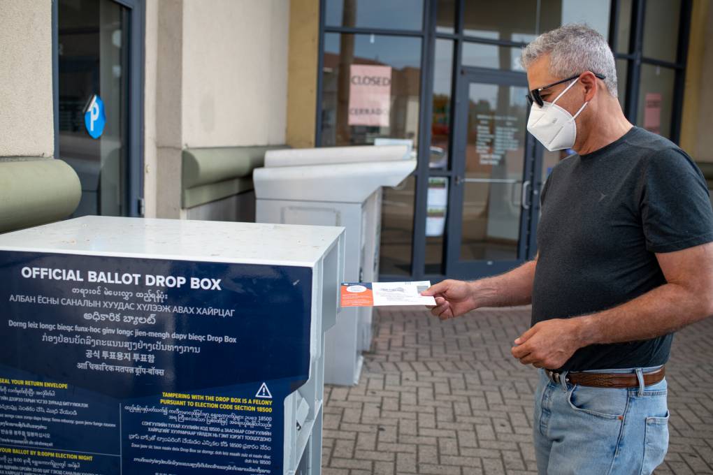 A man in a T-shirt and mask slides a ballot into a drop box.