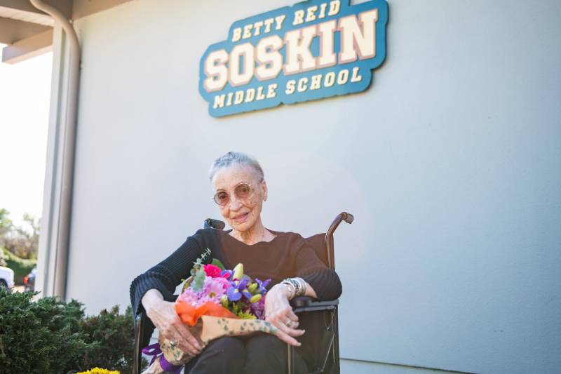 Betty Reid Soskin, smiling, sits in a wheelchair and holds a bright bouquet of flowers.