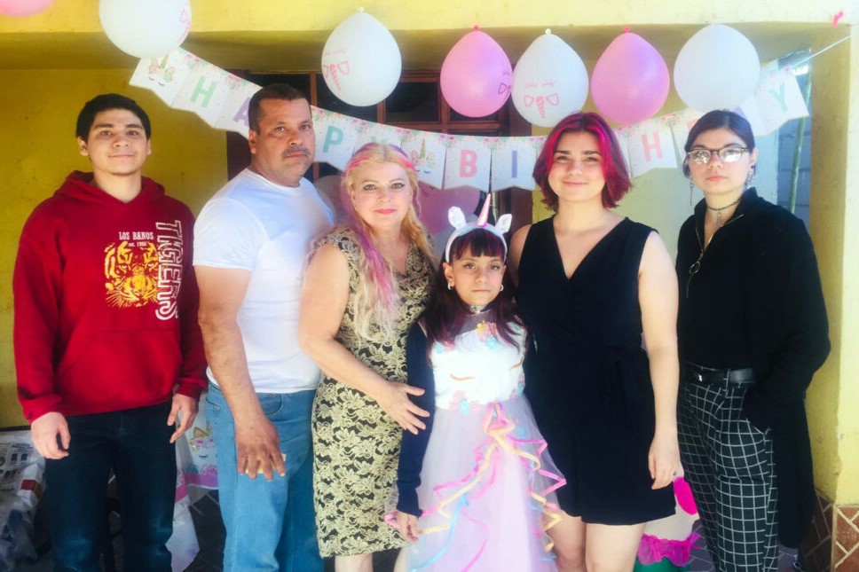 A family stands in a line for a picture amid pink and white birthday decorations. The girl in the middle wears a unicorn outfit.