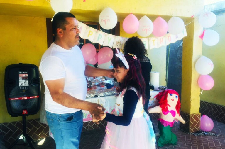 A man in a white T-shirt and jeans holds hands with an 11-year-old girl, amid pink and white balloons and decorations.