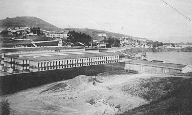 Black-and-white photo of a dirt patch with buildings and a hill behind.