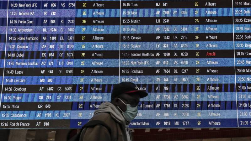 A person wearing a mask and a baseball cap walks in front of an airport departures board.