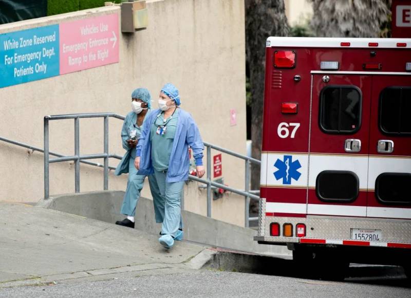 Two nurses in blue scrubs walk up a steep sidewalk outside the ER beside an ambulance parked on the street.