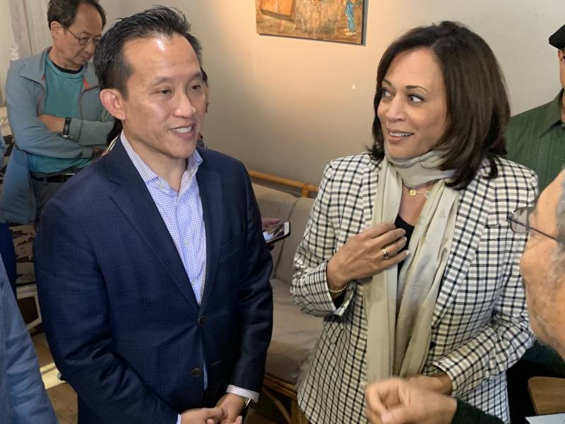 Kamala Harris, in a suit jacket and scarf, looks on as David Chiu, in a suit jacket and no tie, speaks in casual conversation indoors.