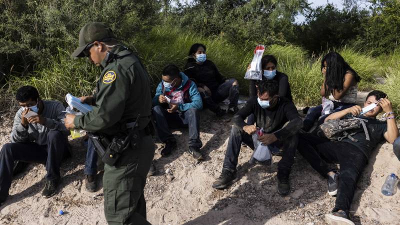 A U.S. border patrol agent in a green uniform stands over a group of migrants, who rest in the sand holding gallon jugs of water.