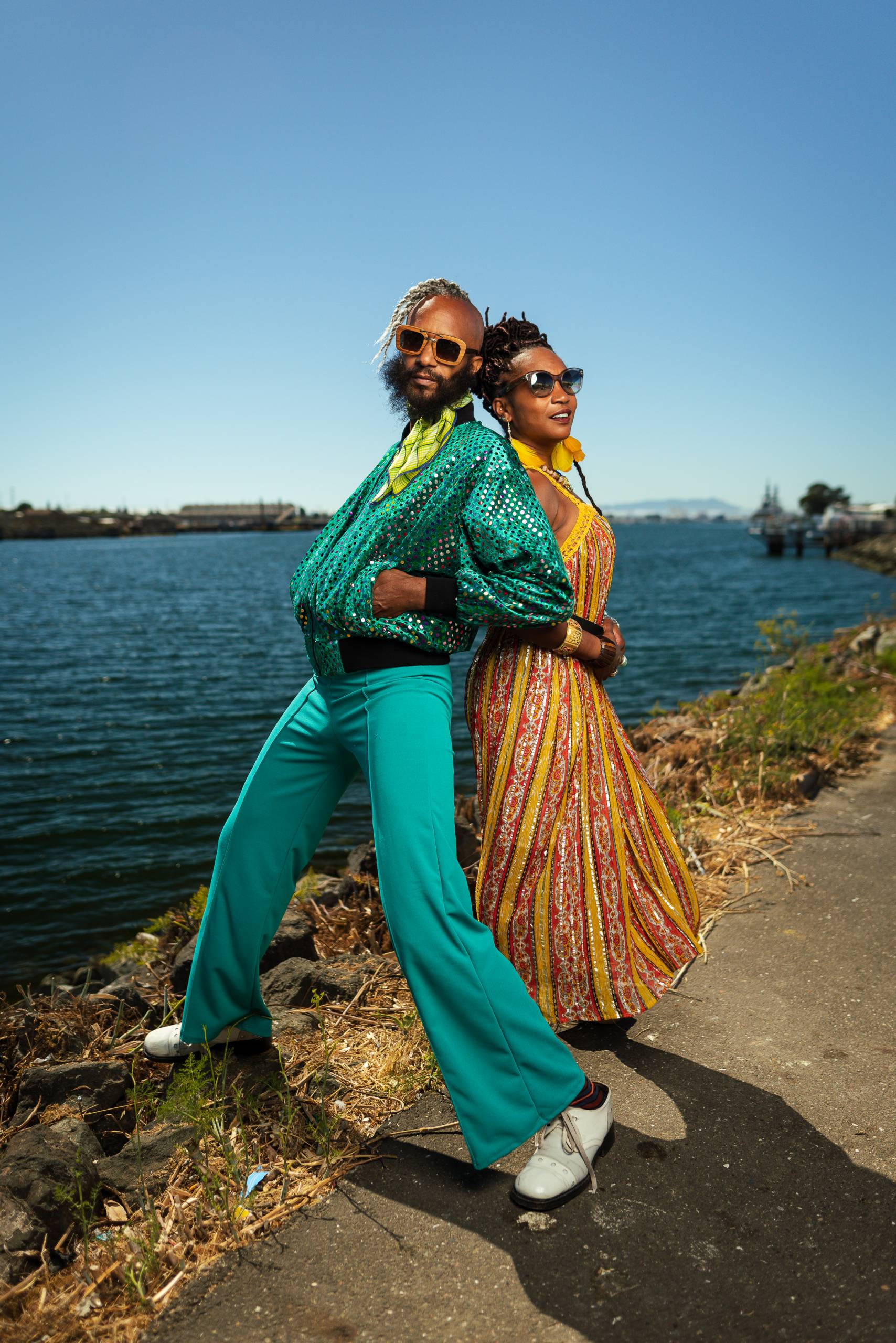 A man and a woman, brightly dressed and wearing sunglasses, stand back-to-back looking at the camera on a path beside a large body of water.