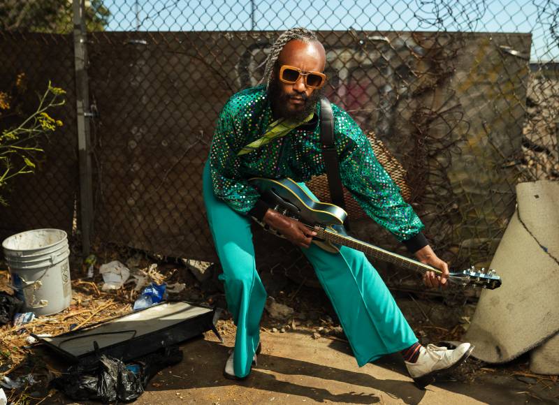 A man in a shiny, teal outfit and sunglasses crouches as he plays a green guitar.