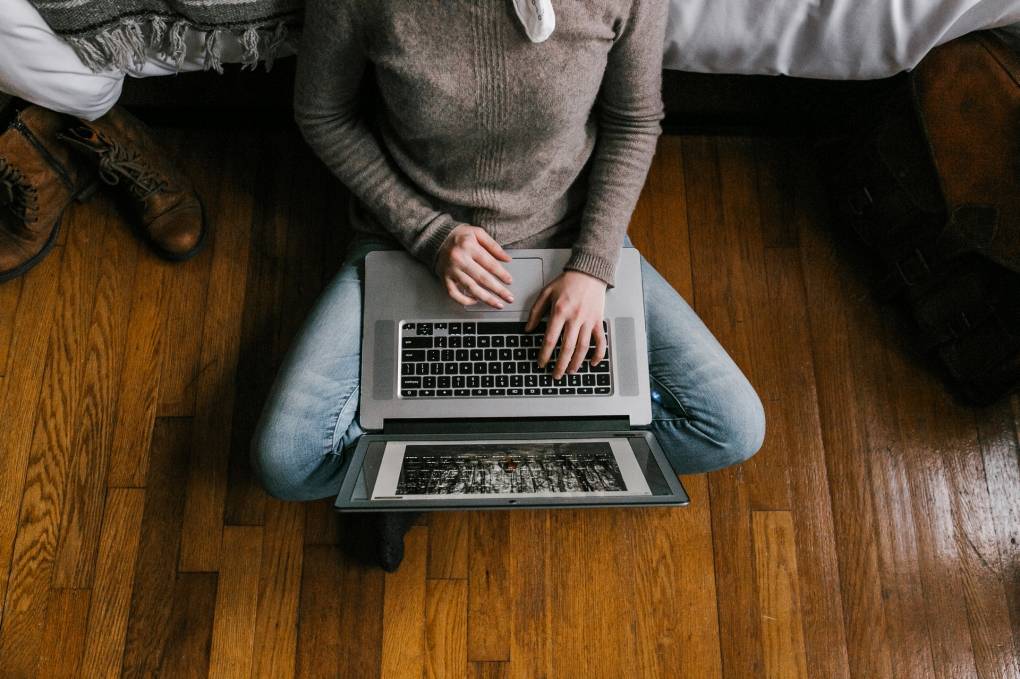A person in a gray sweater sits cross-legged on hardwood floors, using a laptop computer