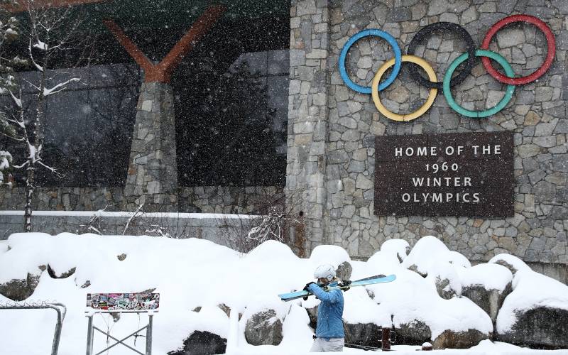 A skier in a bright blue ski outfit holding skis walks in the snow in front of a sign with the Olympic rings