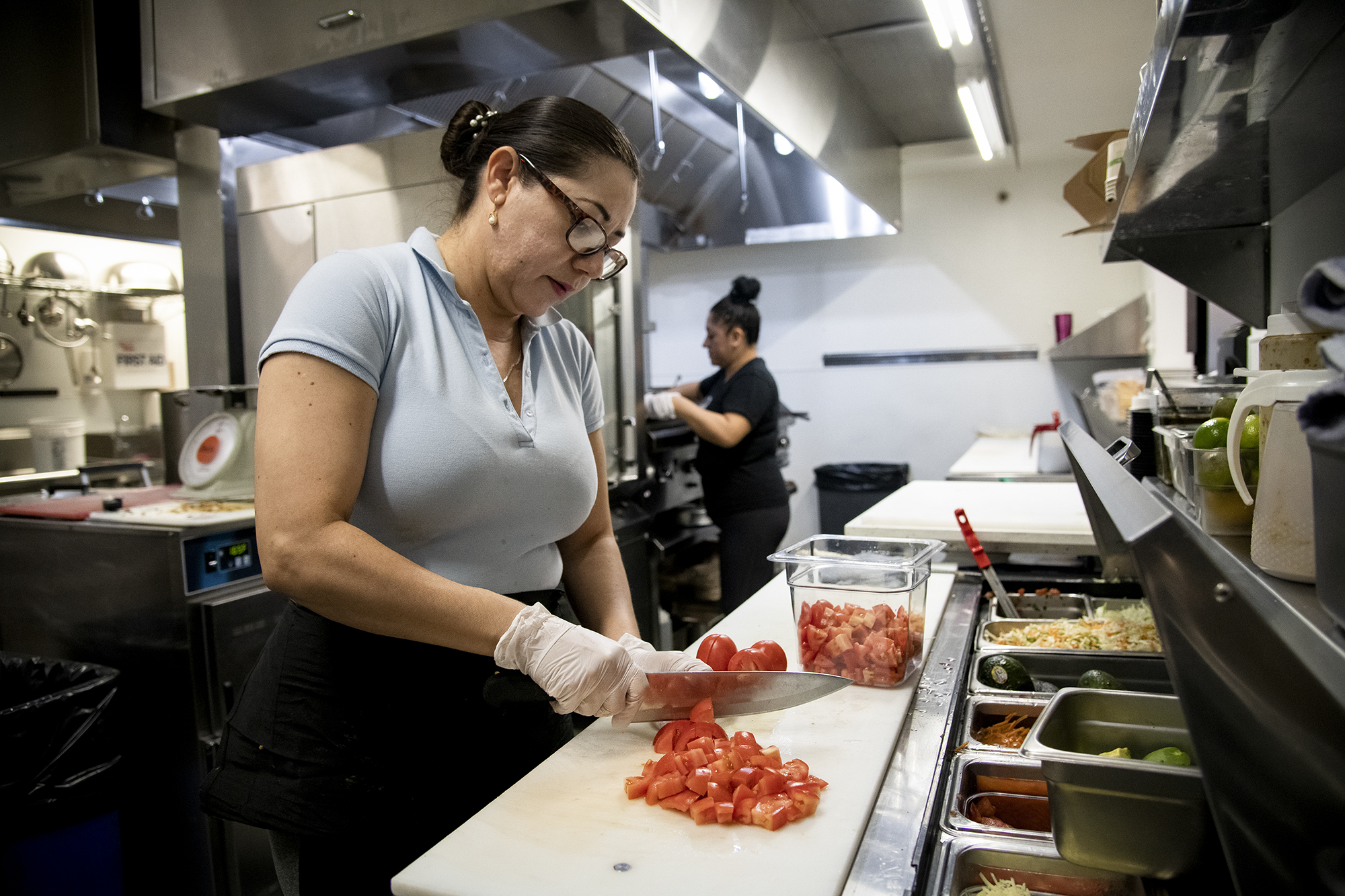 A woman with her hair back and wearing gloves chops tomatoes in a restaurant kitchen.