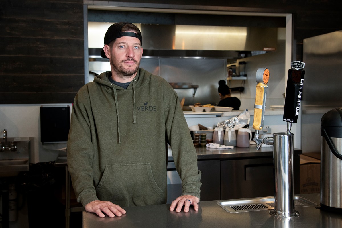 A man in a hooded sweatshirt and a baseball hat on backward stands behind a restaurant counter looking evenly at the camera.