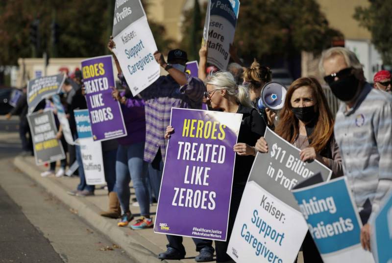 A line of workers on the edge of a sidewalk holding signs that say, for example, "Heroes Treated Like Zeroes."