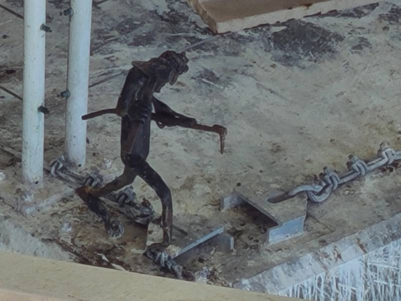 An angular dark metal figurine with legs and arms, holding tools, its feet affixed to the cement below it.