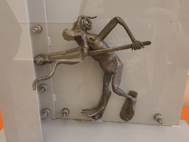 A light-colored metal figurine with two arms and two legs, holding a very long wrench to turn a bolt.