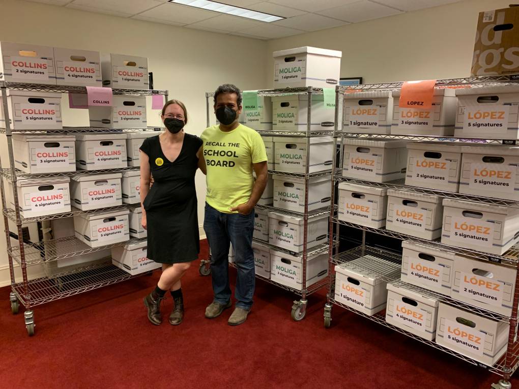 A man and a woman, smiling and arm in arm, stand at the corner of two walls stacked with election filing boxes.