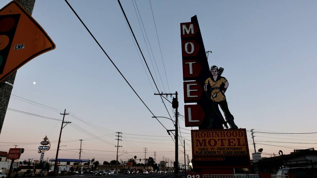 A tall, vertical "Motel" sign with a cowgirl leaning against it, against a sunset and along telephone lines.