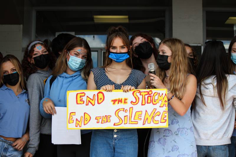 High school students with long hair and wearing face masks hold a sign saying "end the stigma, end the silence."
