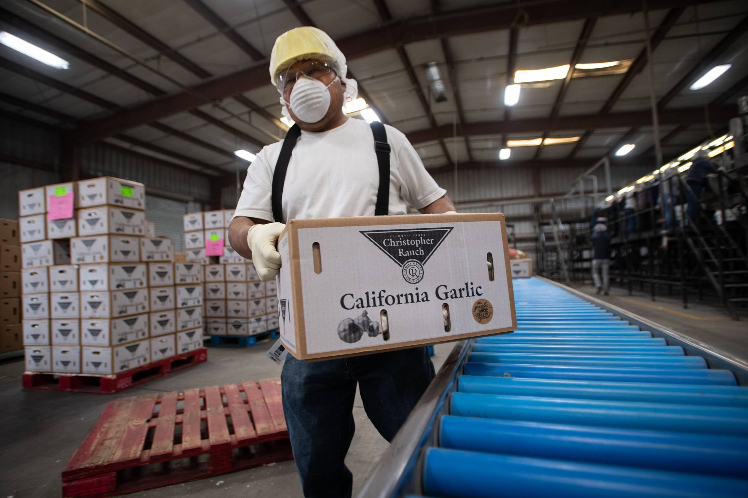 A worker wears a helmet and a face mask as they carry a heavy cardboard box through a factory.