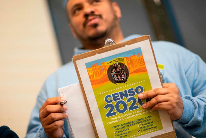 A man in a light blue hoodie holds a clipboard with U.S. census information in Spanish on it.