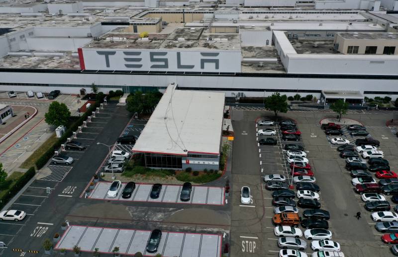 An aerial view of a long, flat building, a massive Tesla sign, and a massive parking lot.