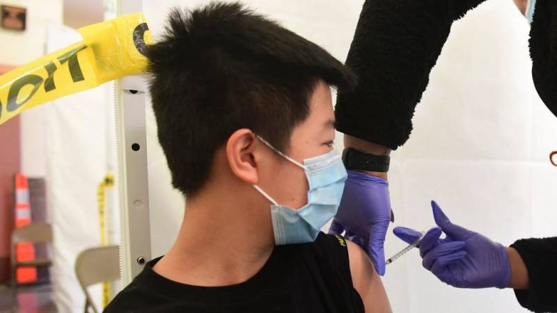 A boy wearing a mask and sitting along a wall with student work displayed looks at his shoulder as someone wearing purple gloves injects his shoulder.