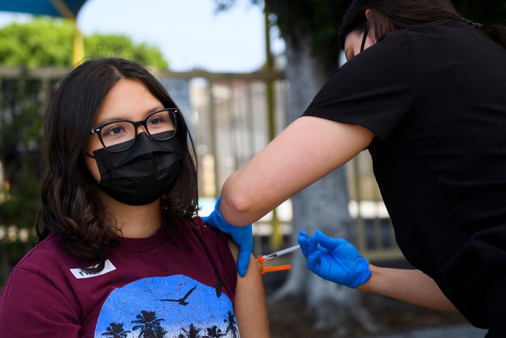 A teen girl wearing glasses and a mask, sleeve rolled up, looks past a nurse with blue gloves who grips her shoulder with one hand and injects her with the other.