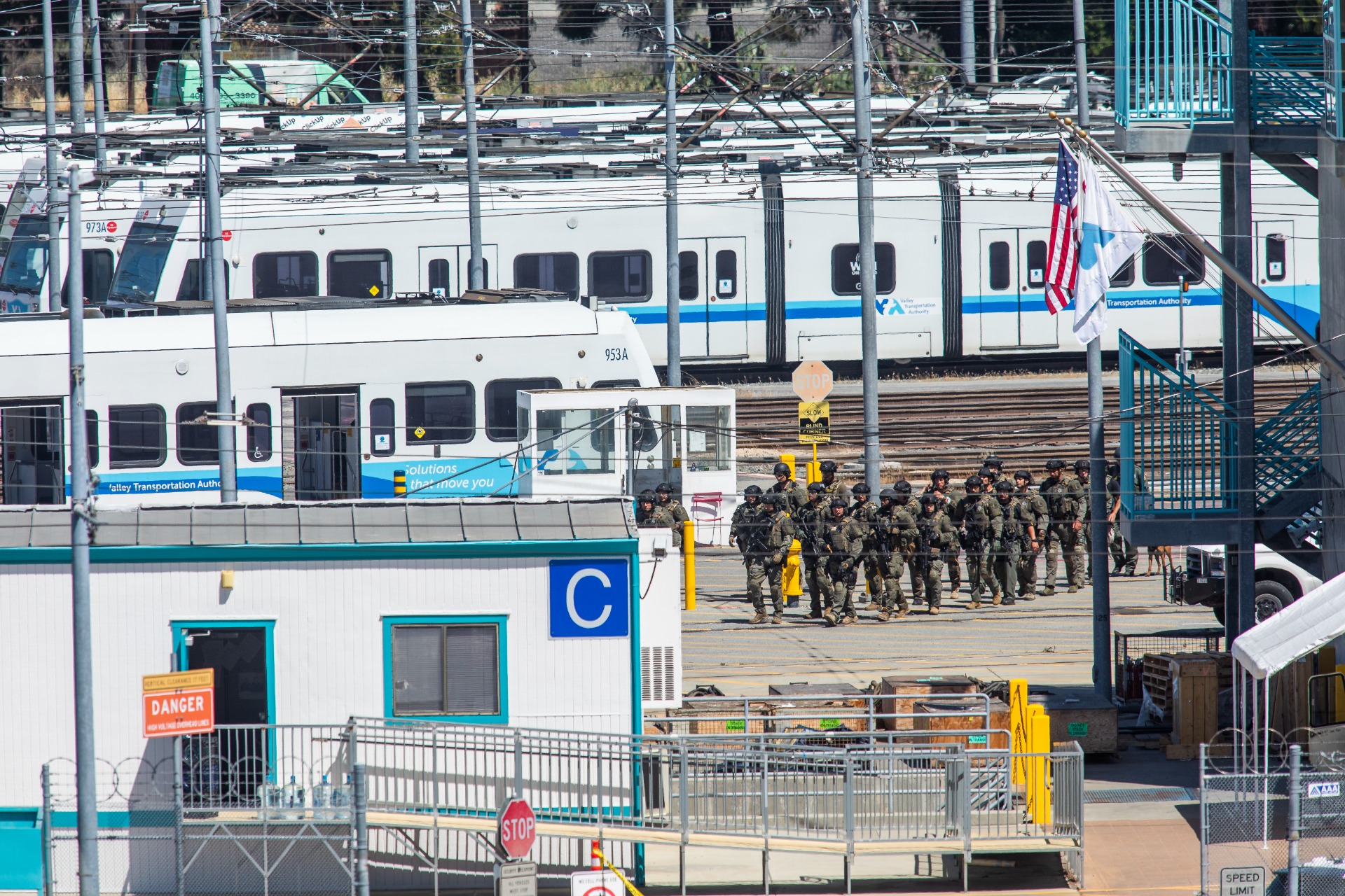 A faraway shot of a rail yard with a line of trains, as a small crowd of people in tactical gear walk toward the camera. 