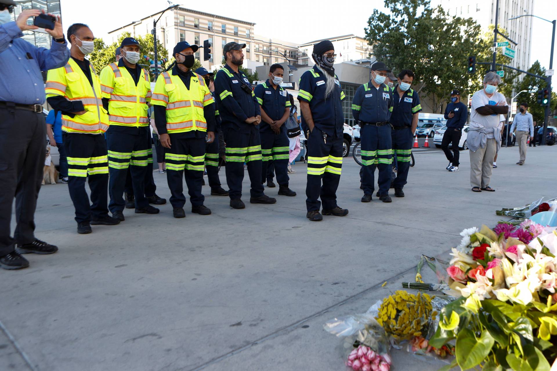 A line of people in uniform -- neon yellow jackets and dark blue pants with reflective stripes at the knees -- stand before a collection of flowers.