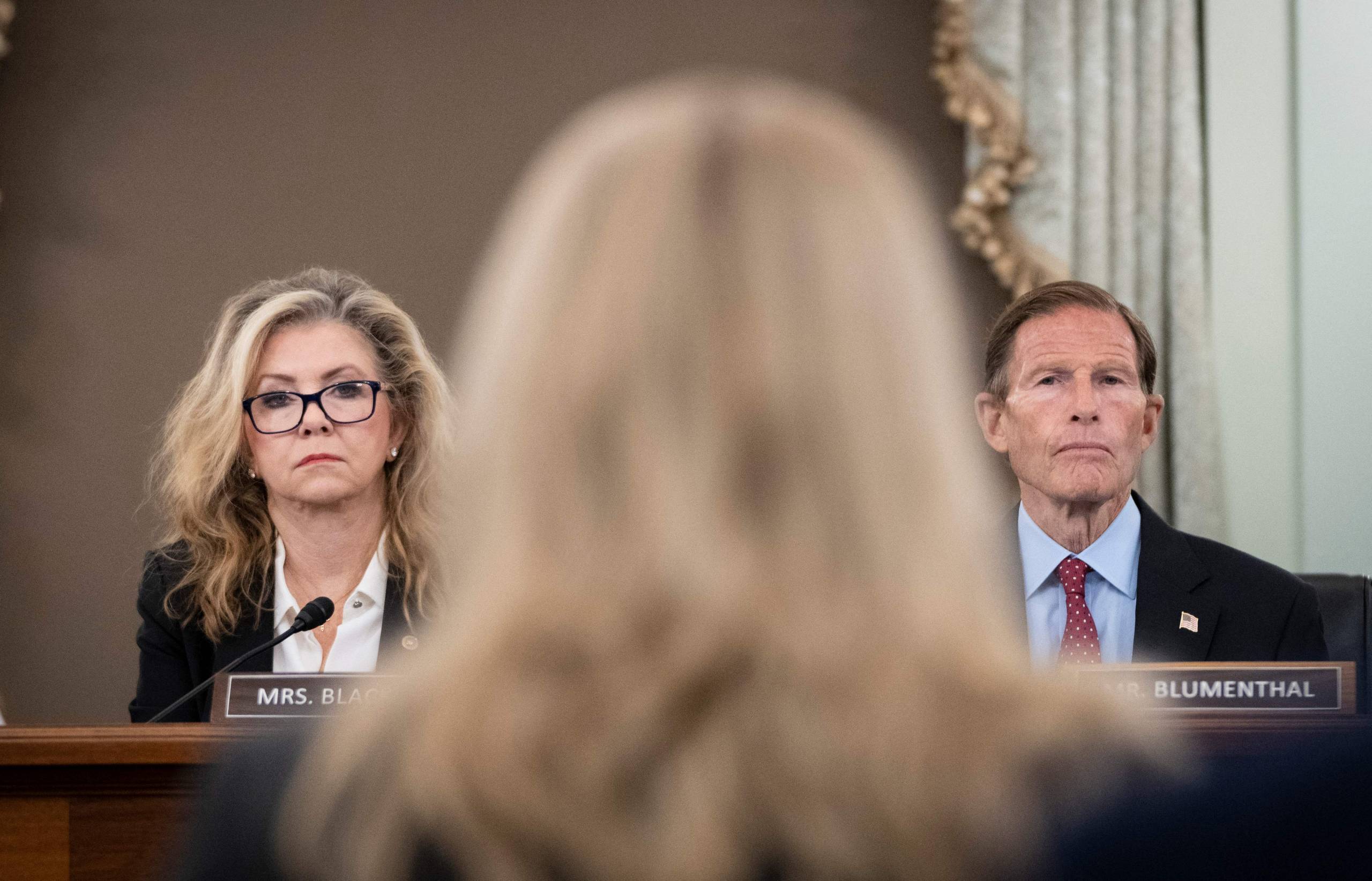 In the foreground is the back of Frances Haugen's out-of-focus head, and seated beyond her, and in focus, are Senators Marsha Blackburn and Richard Blumenthal on either side of her head.