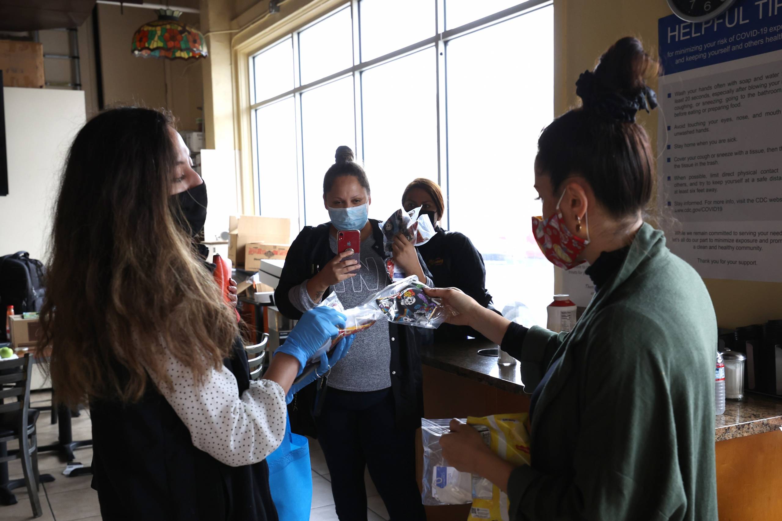 Three women wearing masks gathers inside a cafe. One hands another a set of face masks while the third records the scene on their phone.