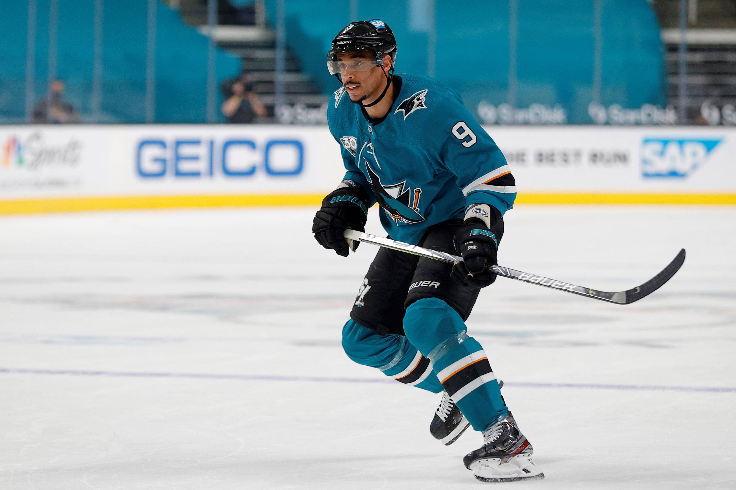 Evander Kane skates on the ice rink and wears a Sharks jersey and holds his hockey stick.