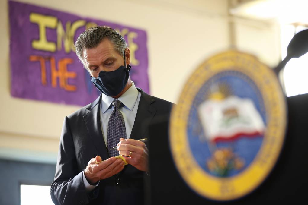 Gavin Newsom, wearing a black mask and standing beyond a podium with the Seal of California, scribbles in a tiny notepad.