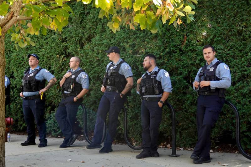 Five police officers in bulletproof vests lean against metal bike racks in front of an ivy-covered wall.