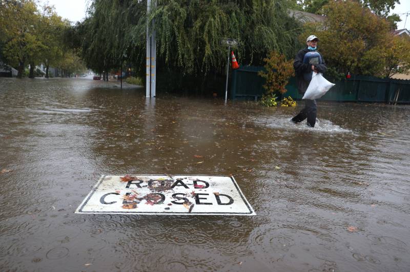 The "Road Closed" sign floats on a sea of brown water. A man holding a plastic bag wades through ankle-deep water beyond it.