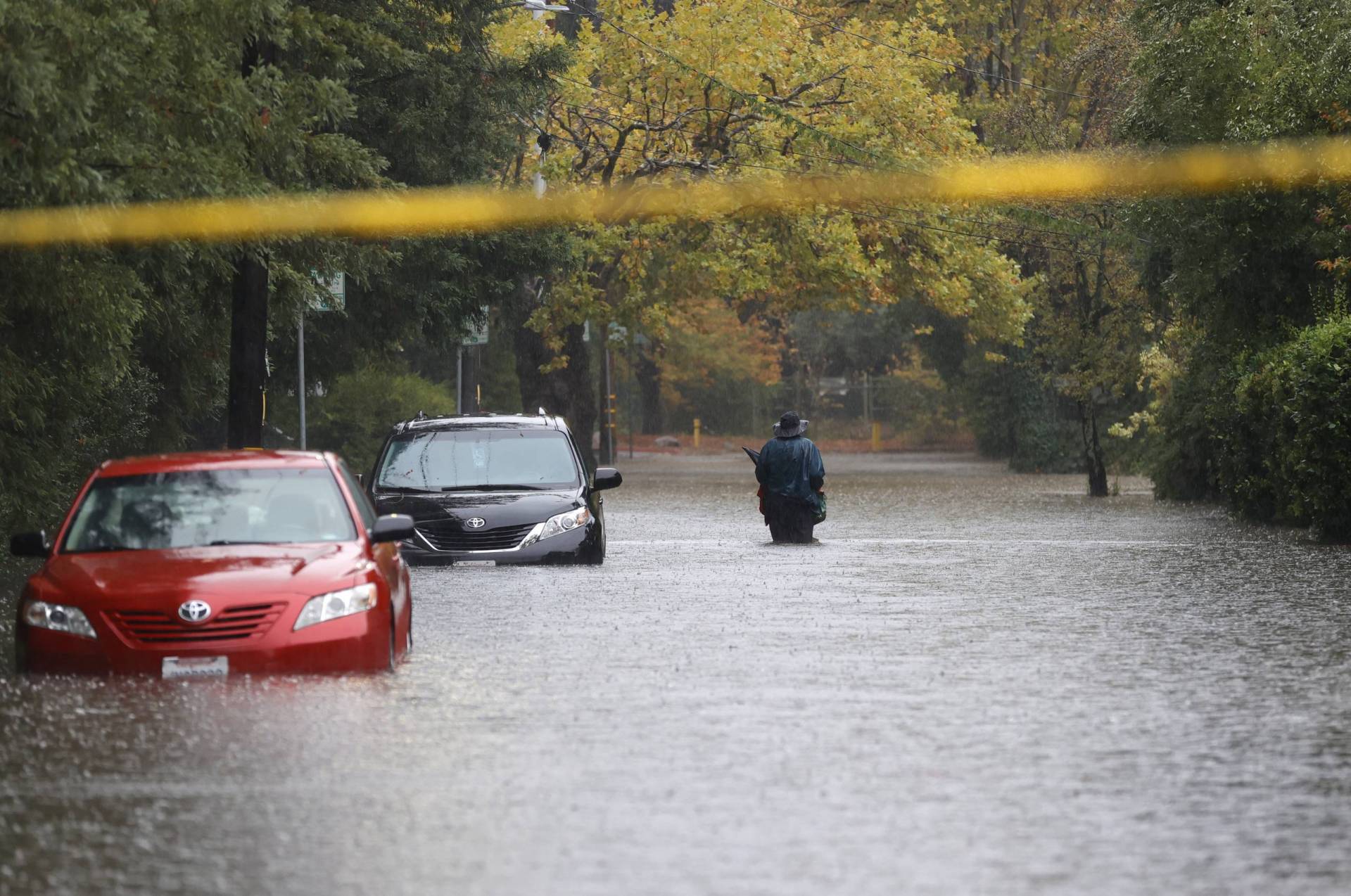 A man wades through water higher than his knees next to two parked cars past their bumpers in water, on a tree-lined street, beyond blurred yellow police tape in the foreground.