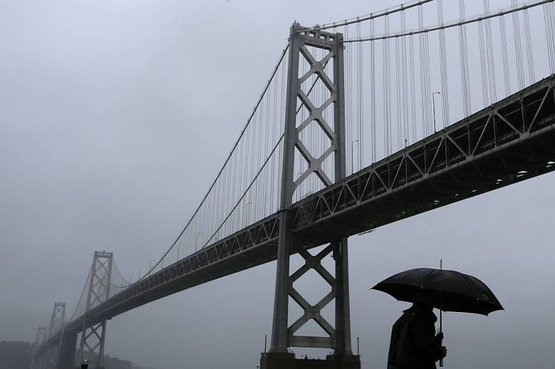 A dark silhouette of a person with an umbrella walking through dense gray fog and rain in front of the Bay Bridge.