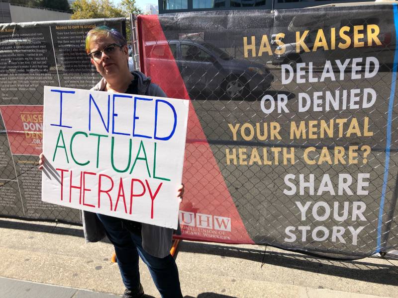 A woman holds a sign saying "I Need Actual Therapy" in front of a sign on a chain link fence reading, "Has Kaiser Delayed or Denied Your Mental Health Care? Share Your Story."