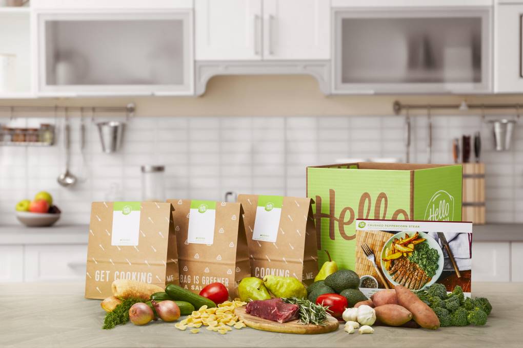 A company photo in a white kitchen of four brown paper Hello Fresh bags with a bright green color, fresh vegetables arrayed on the counter in front of them.