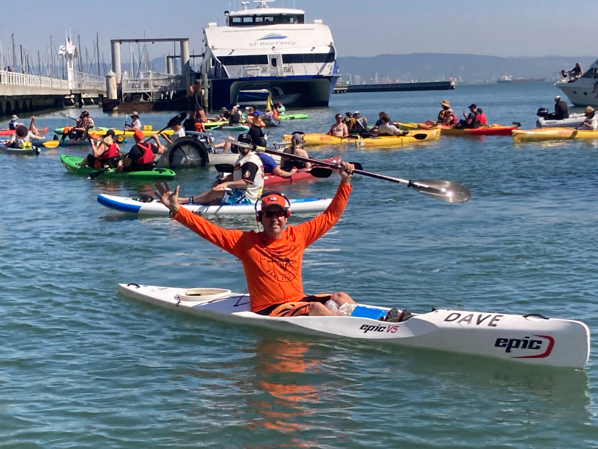 A man in an orange shirt and hat and wearing headphones holds his oar above his head triumphantly while sitting atop a white kayak in San Francisco Bay, surrounded by other boaters.