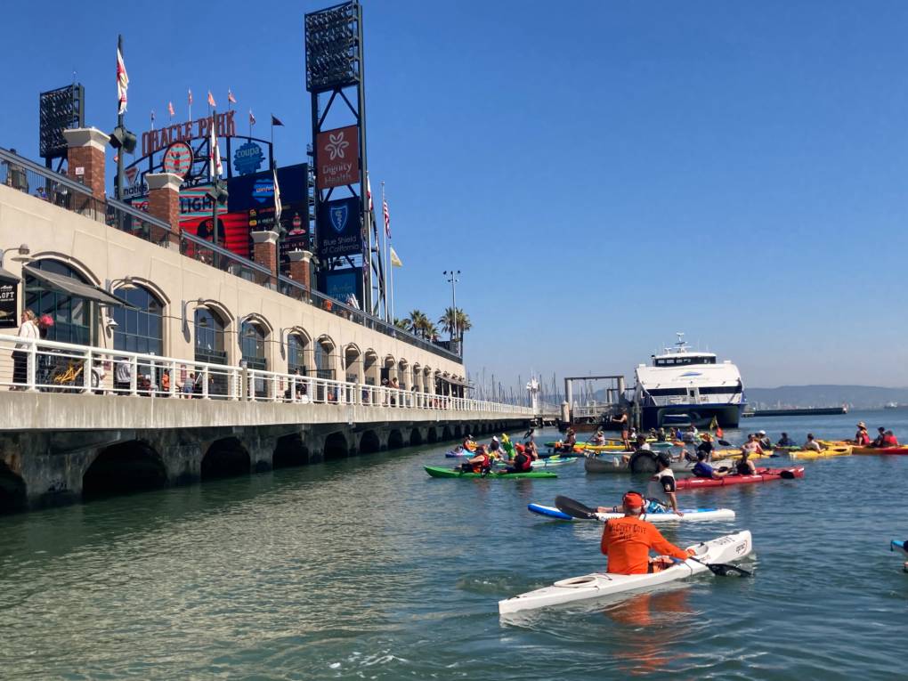 Man dressed in orange on a white kayak floats in the water beyond the facade of the ballpark.