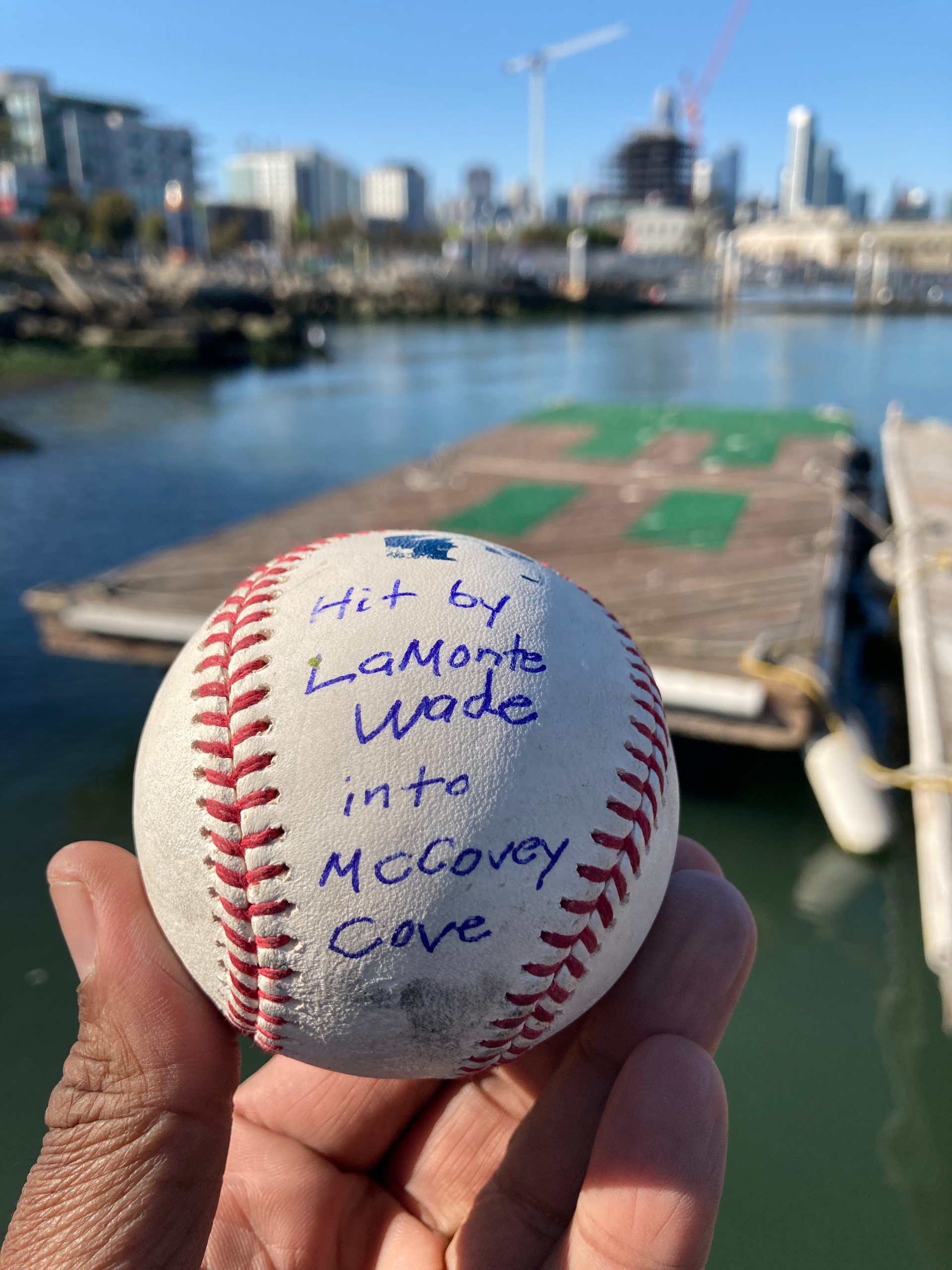 A hand holds a baseball with writing on it that says: "Hit by LaMonte Wade into McCovey Cove." Blurry city landscape with water beyond it.