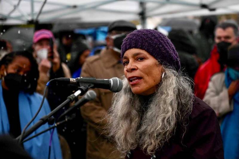 A woman with long gray hair and a purple knitted cap speaks into a microphone, amid people holding umbrellas.