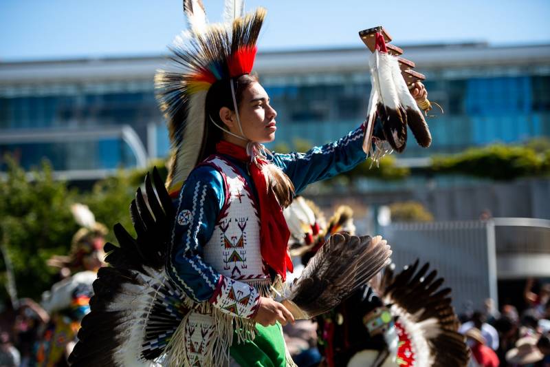 A dancer in a red, blue, and white costume, including a headdress and a feathered tail, holds feathers in both hands and faces the sun.